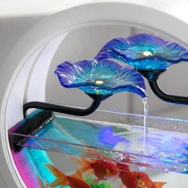 A close-up view of the blue glass flowers at the top of an aquarium waterfall air purifier. Water can be seen flowing from one flower to the next and flowing back into the tank. There are goldfish inside the fish tank hanging around near the waterfall.