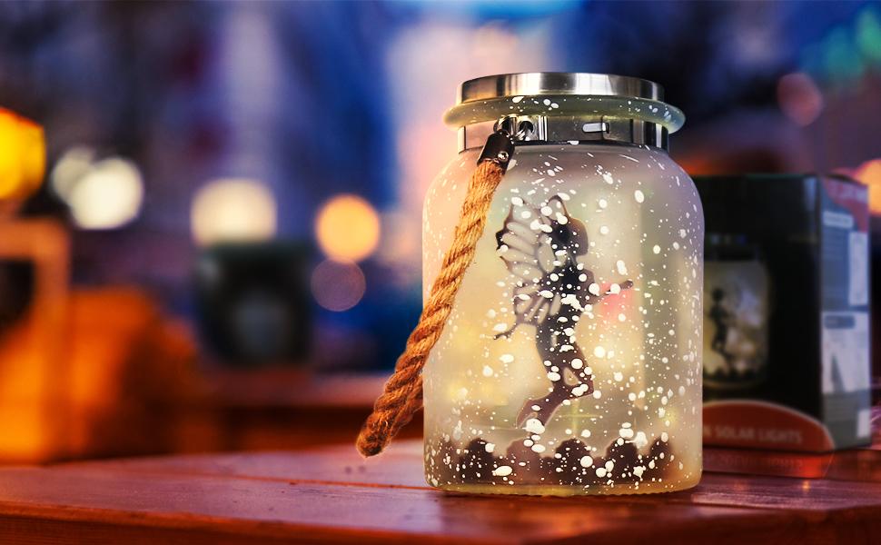A solar mason jar lit up with the silhouette of a fairy inside resting on a table.