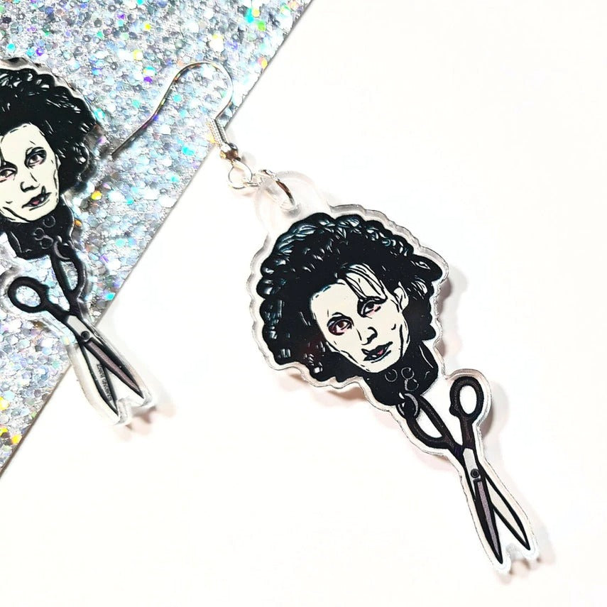 Close up view of a pair of earrrings inspired by the 90's movie Edward Scissorhands. They feature Edward Scissorhands head with a pair of scissors handing off the end. These are resting on a white and silver glitter background.
