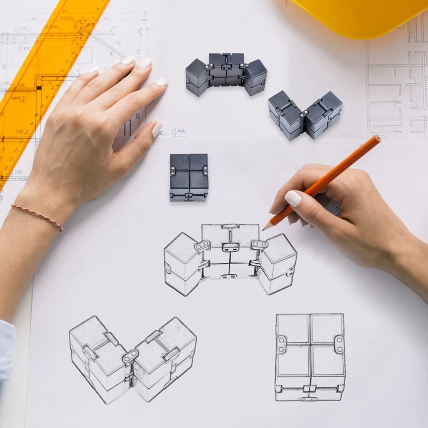 A pair of hands is holding a pencil and drawing a fidget cube on paper. Next to the drawing is an actual fidget cube.