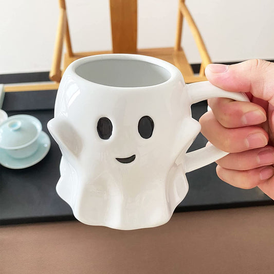 A hand holding a white ceramic ghost shaped coffee mug with a smiling and happy expression.
