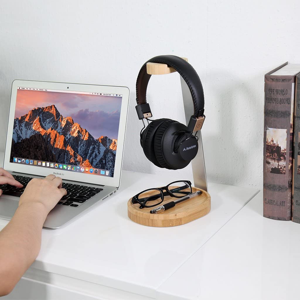 A black pair of headphones on a timber stand. Below the stand there is a cable-tidy which is holding a pair of glasses and a pen. A pair of hands are typing on a keyboard next to the stand.