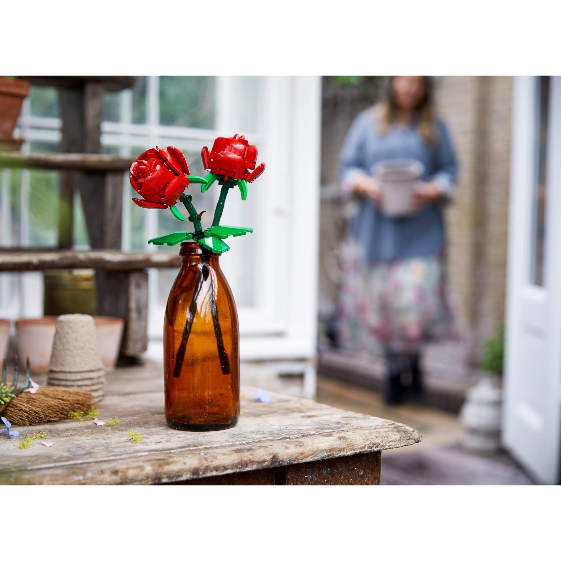 Two Lego red roses in a brown bottle on an old rustic wooden table. There is a woman in the background.