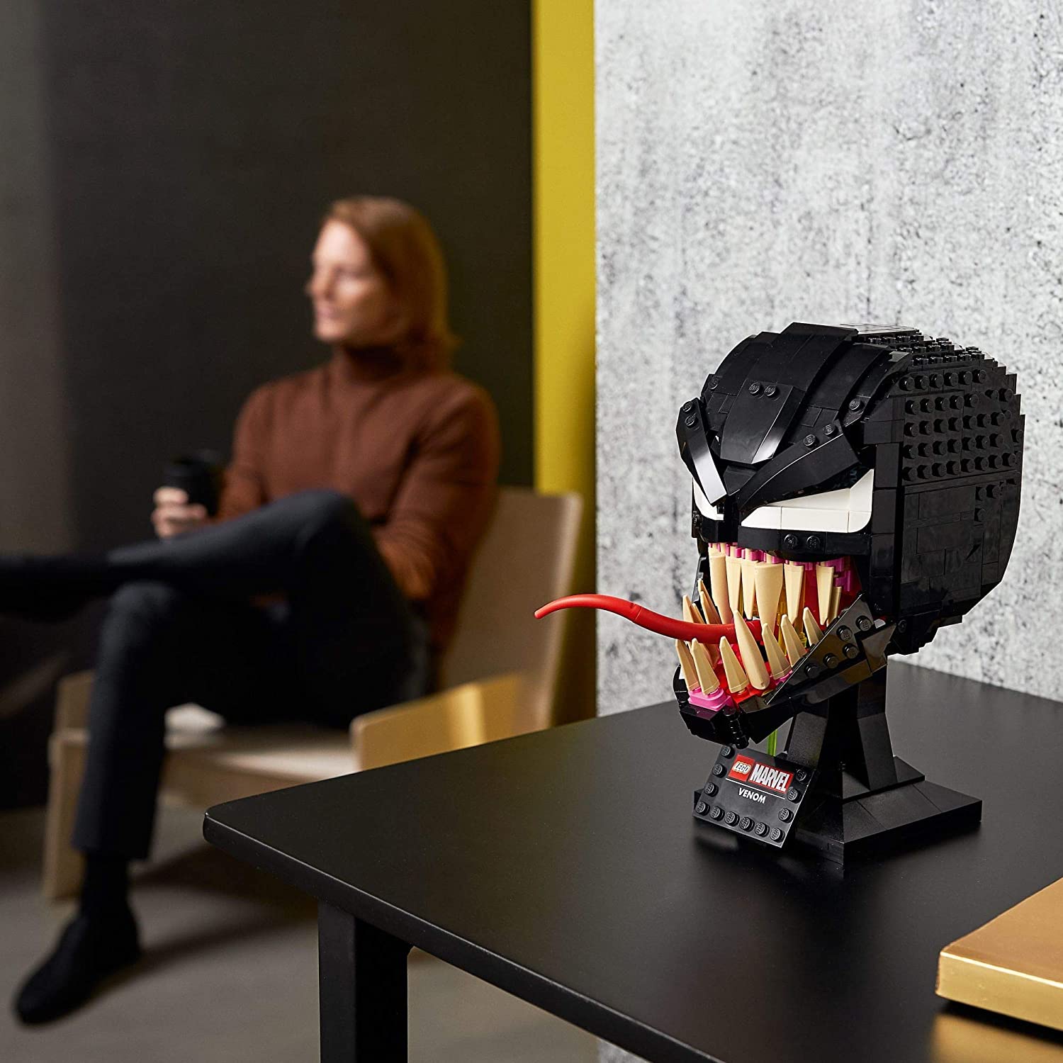 A fully completed model of Venom made from Lego sitting on a black table. In the background a person is sitting in a chair with a cup of coffee.