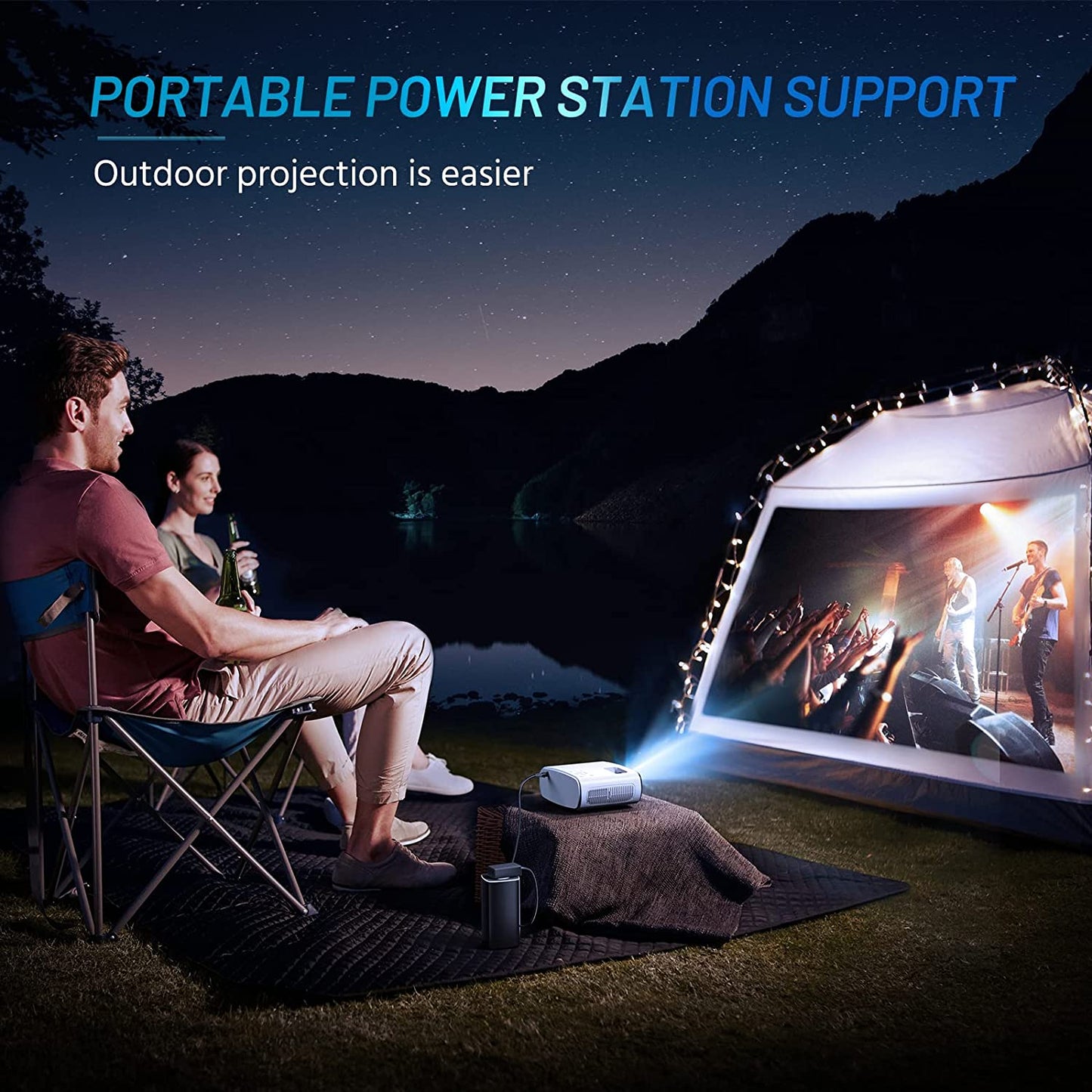 A man and woman are sitting outdoors in front of a tent. They have a mini portable projector displaying a movie onto the side of a tent.