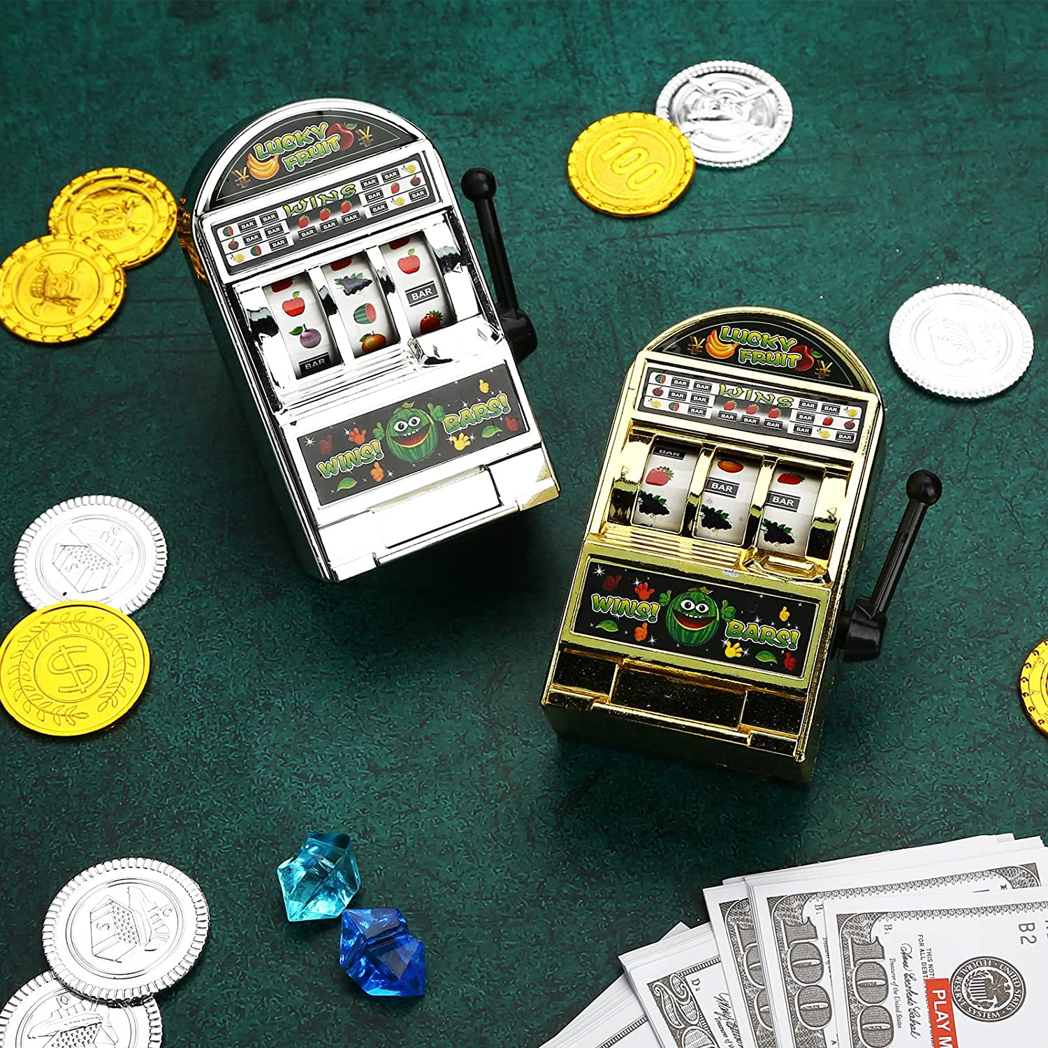 One gold and one silver mini slot machine toys laying down on green velvet with casino coin chips scattered around them.