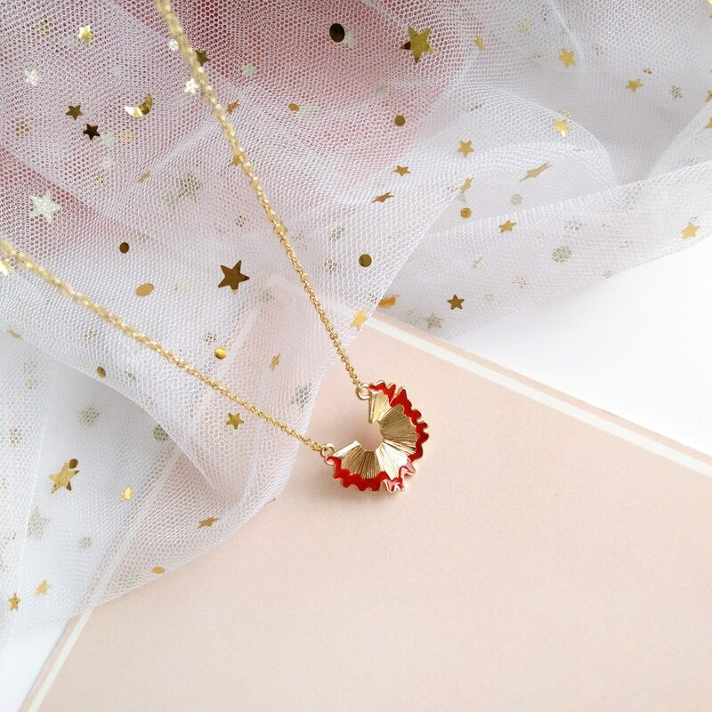A gold pencil shaving necklace which features a gold chain and a gold pendant in the shape of a red pencil shaving.