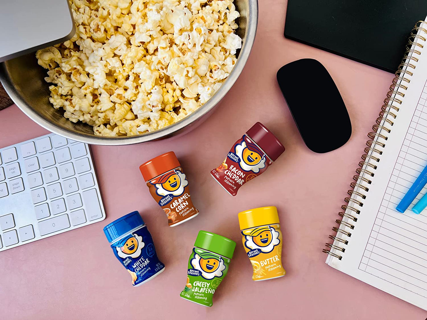 Five different flavoured popcorn seasoning jars on a table with a large bowl of popcorn nearby.