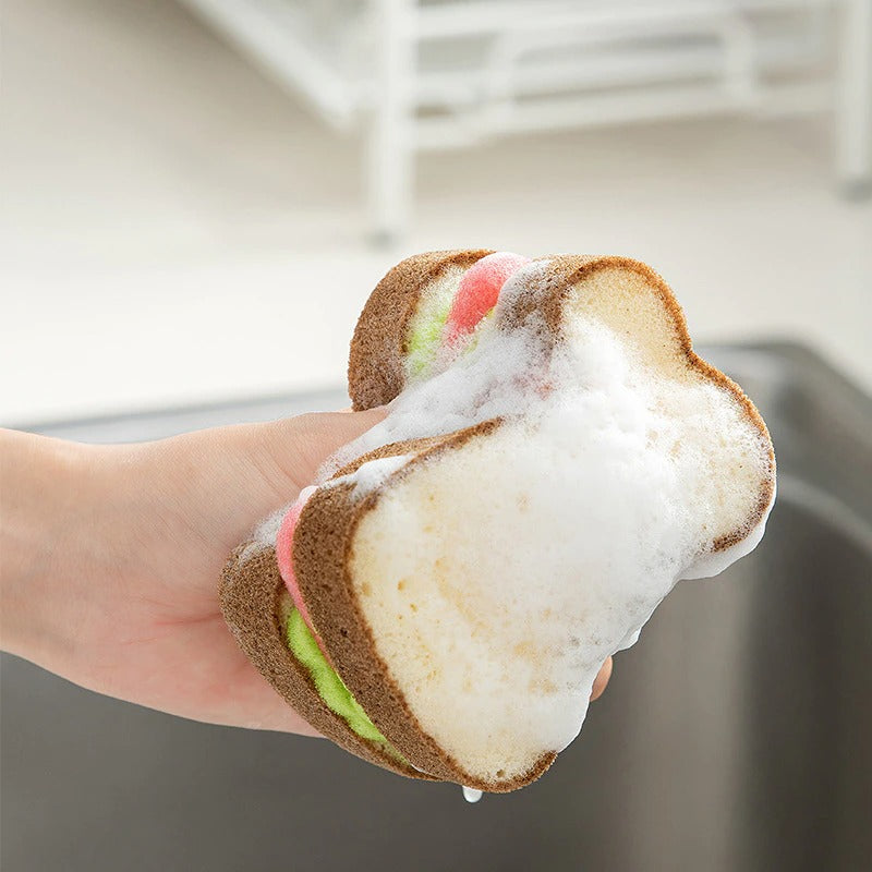 A hand holding a kitchen sponge which looks like a sandwich with two slices of bread and filling. The hand is squeezing the sponge releasing soapy suds.