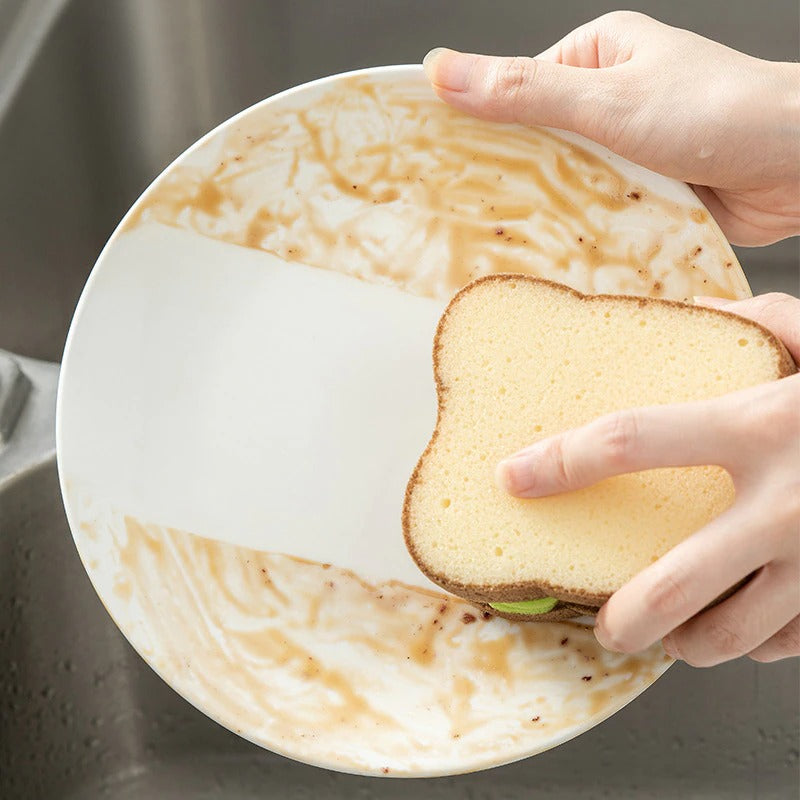 A hand is using a bread sponge to clean a plate in a sink. The sponge is shaped and colored like a slice of bread.