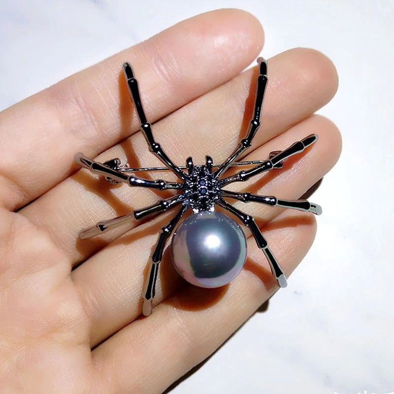 A black spider brooch with small faux black gems and a simulated black pearl. The brooch is on the fingertips of a palm showing its size and details closeup.