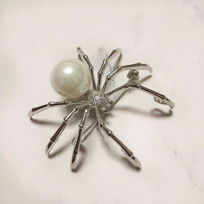 A silver colored spider brooch with a white simulated pearl sitting on a white background.