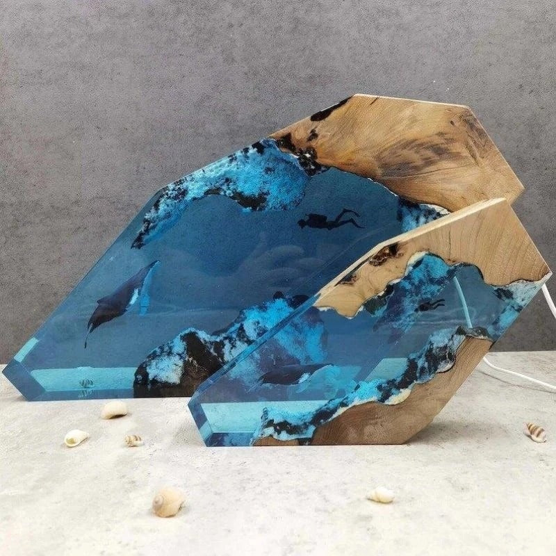 Two desk lamps which look like a slice of underwater life. One lamp is larger than the other showing the two size differences available to buy. The lights are not switched on. There is a diver and a humpback whale swimming toward each other in an underwater cave within the lamp.