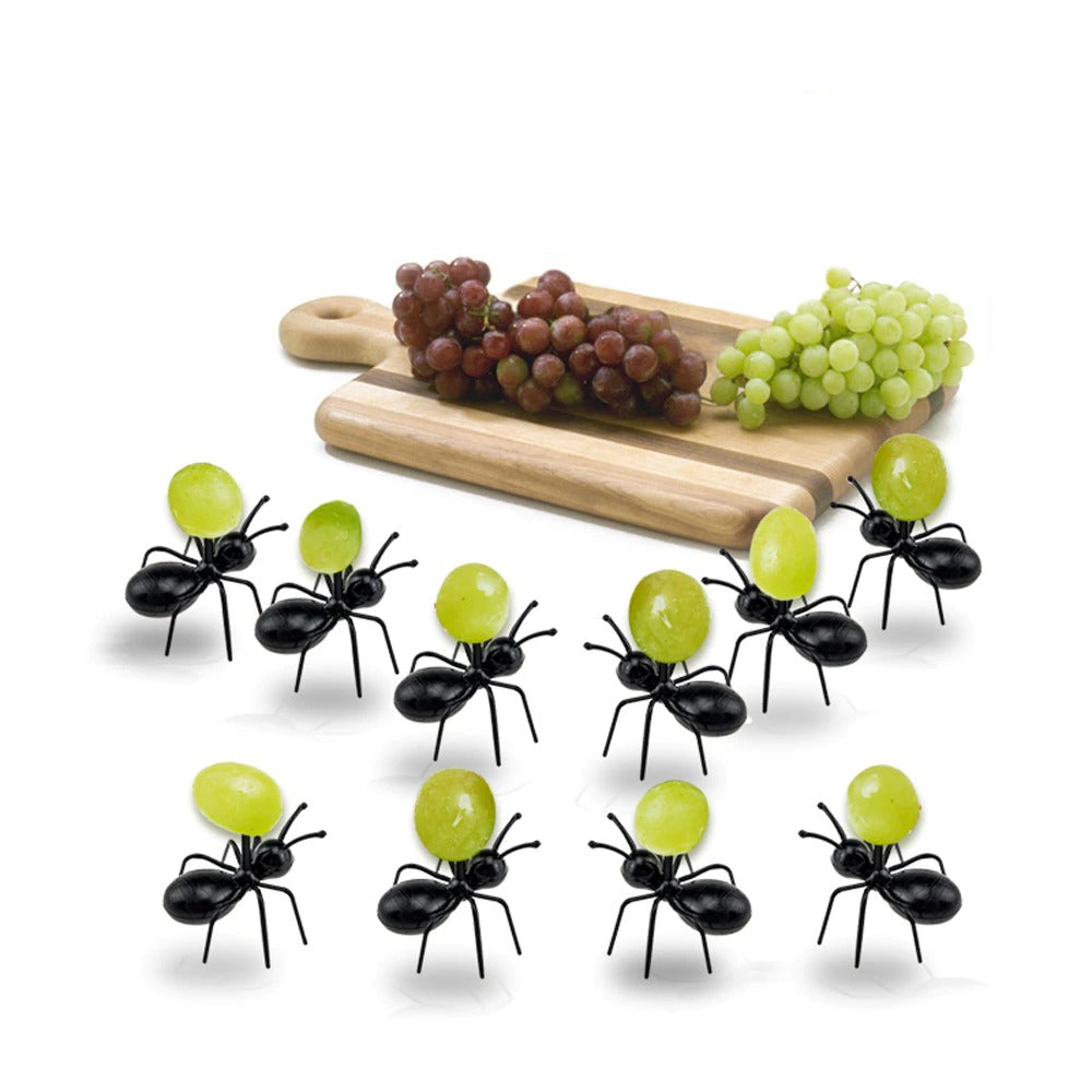 10 ant food picks made from plastic. They are all carrying one green grape each. In the background is a chopping board with a bunch of red and a bunch of green grapes on it.