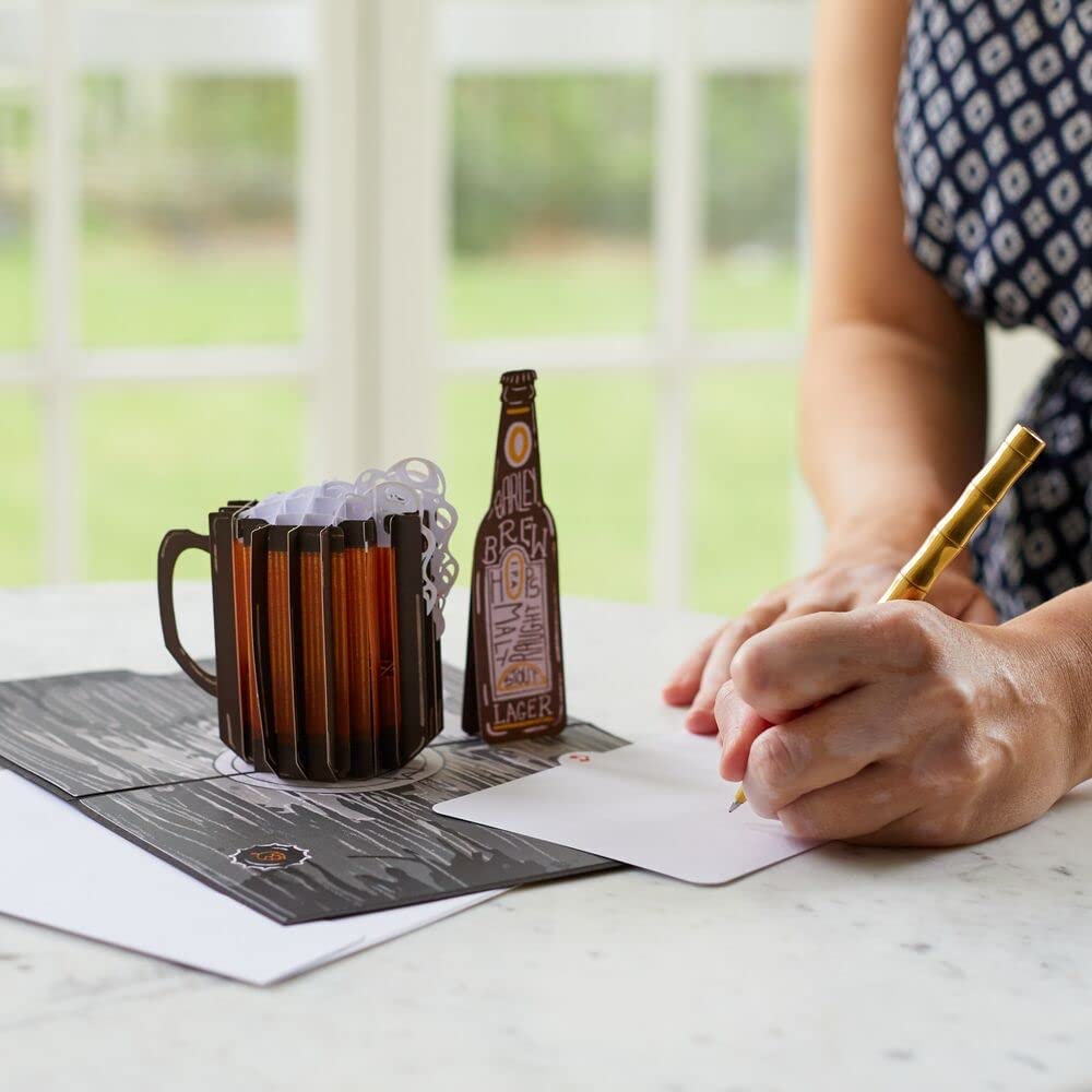 A person is writing inside a 3D pop-up greeting card featuring a beer bottle and a beer stein.