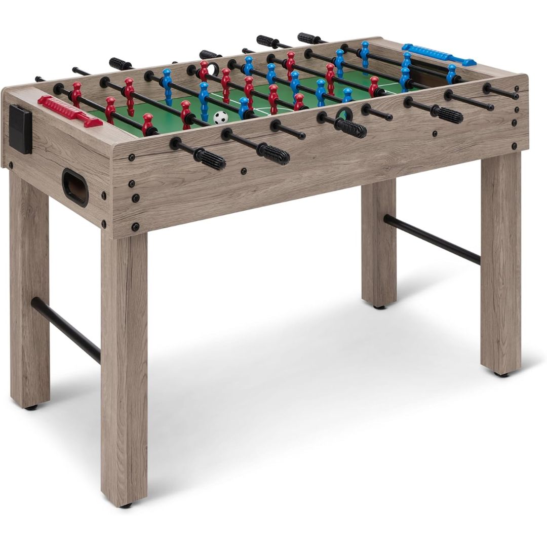 A wooden foosball table with a light gray-brown wood-grain finish, featuring a green playing field, red and blue player figures, black handles, and a white soccer ball on a clean white background.