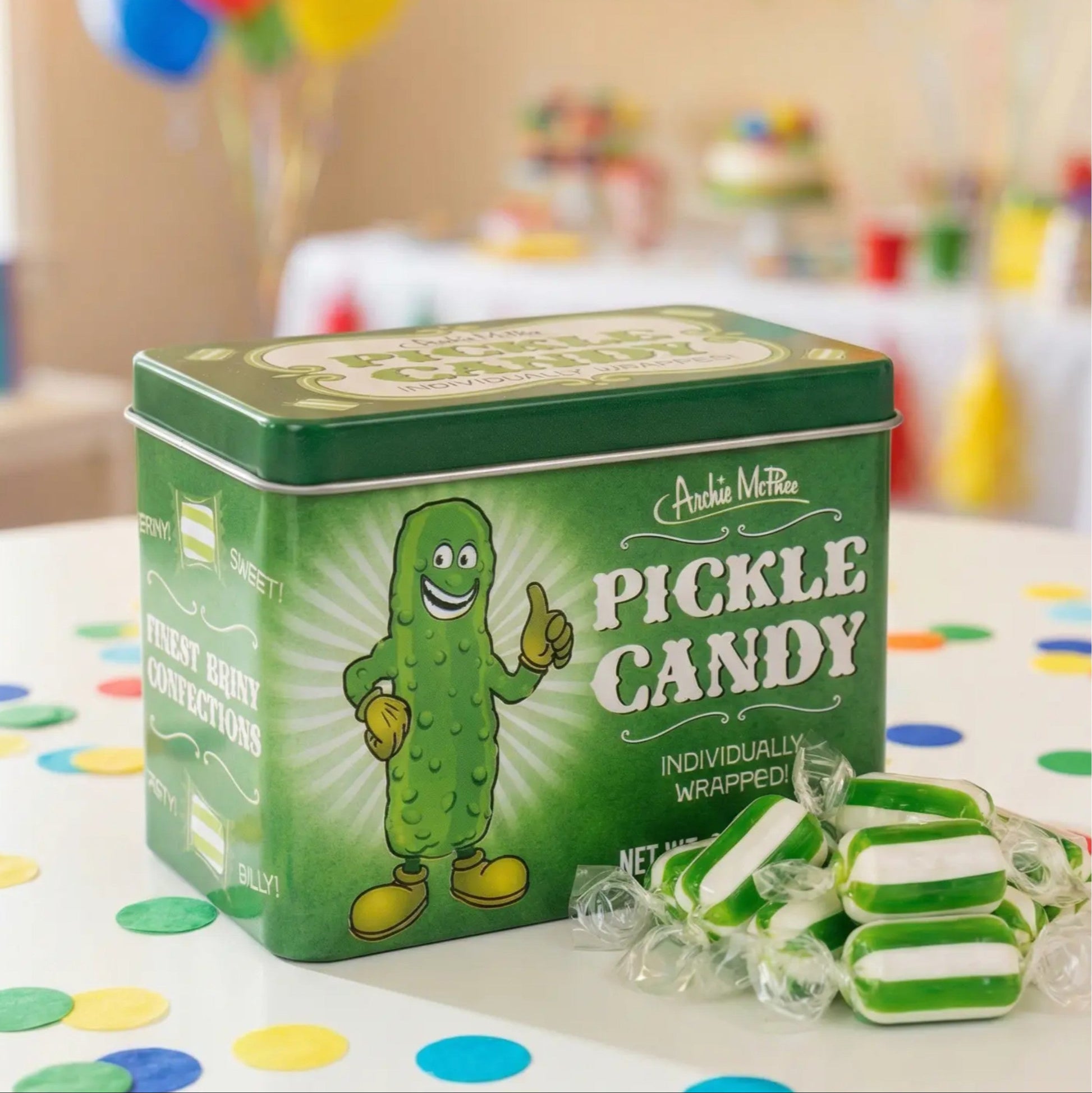 Green tin of pickle candy with a cartoon pickle character on a table with colorful balloons and decorations.