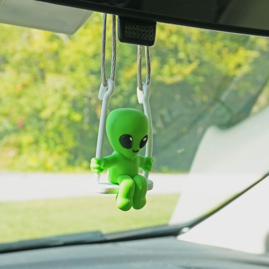 A small green alien figurine sitting on a white swing, hanging from the rearview mirror of a car with a blurred green outdoor background.