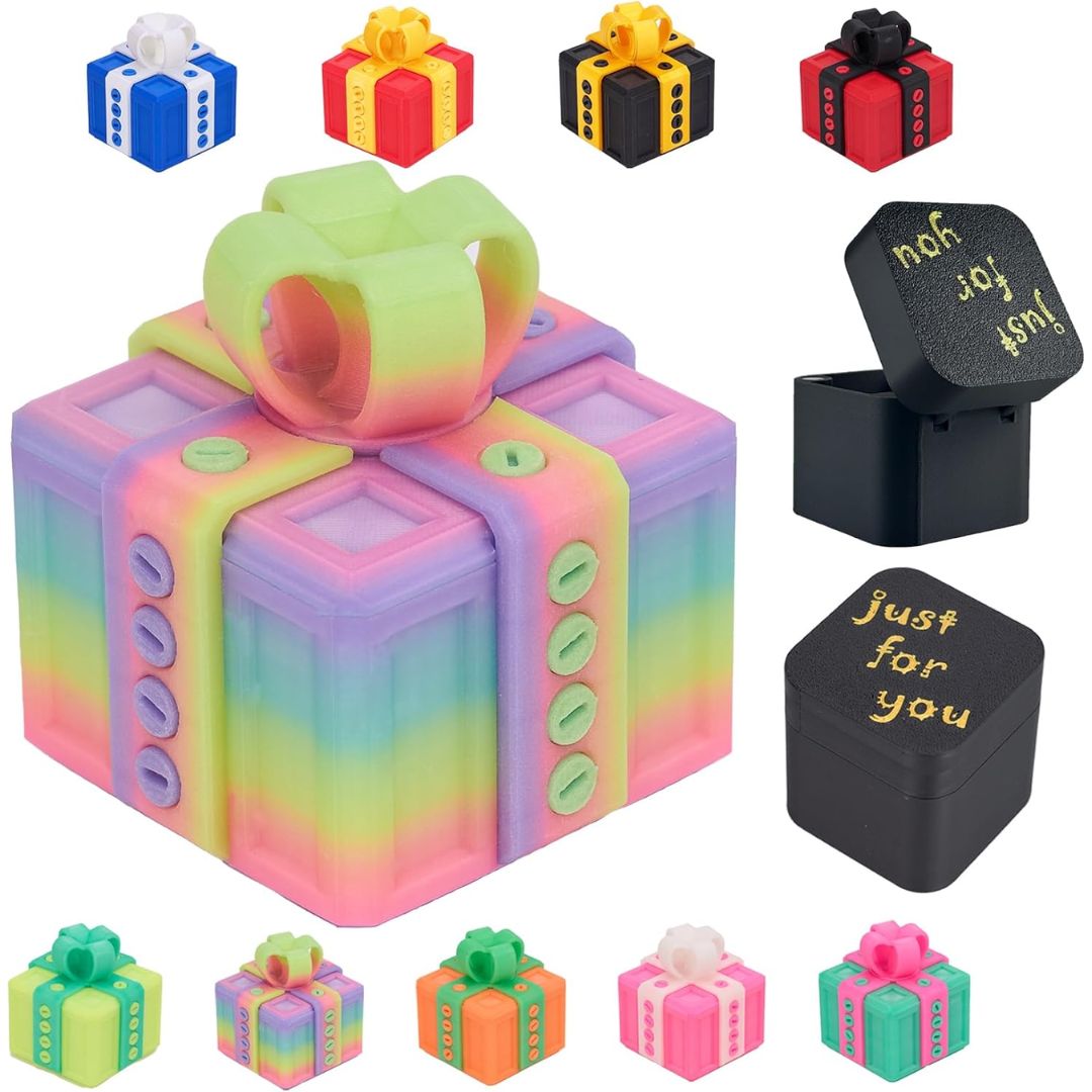 An annoying gift box-shaped ring box with a pastel rainbow gradient and a lime green ring, shown alongside several small gift box ring boxes in different colors and one open black box with gold text. The main product is centered on a plain white background.
