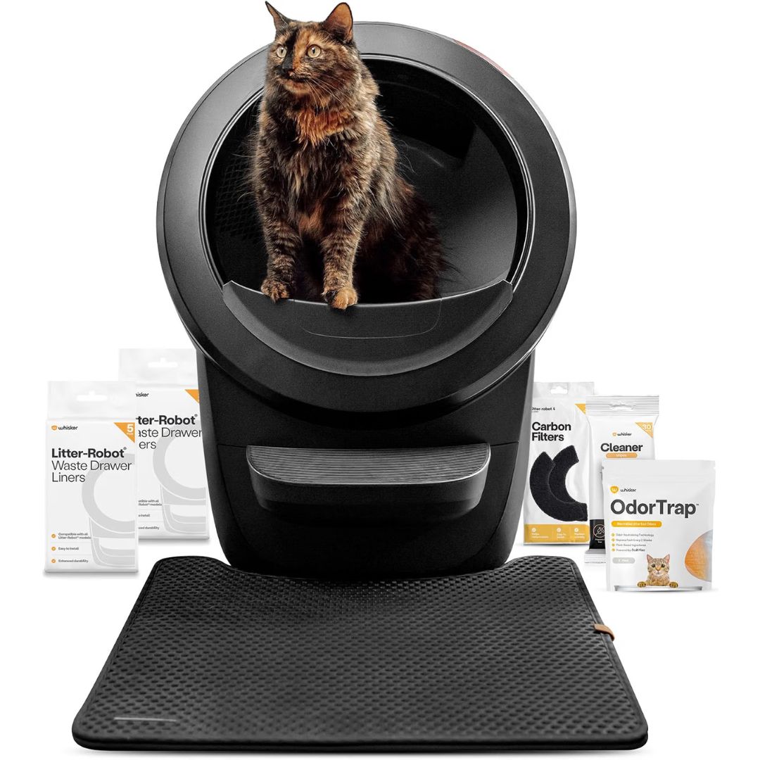 A black automatic self cleaning litter box with a round opening, shown with a brown tabby cat sitting inside. Several product accessory packages are arranged around the base, and a black textured mat lies in front on a white studio background.
