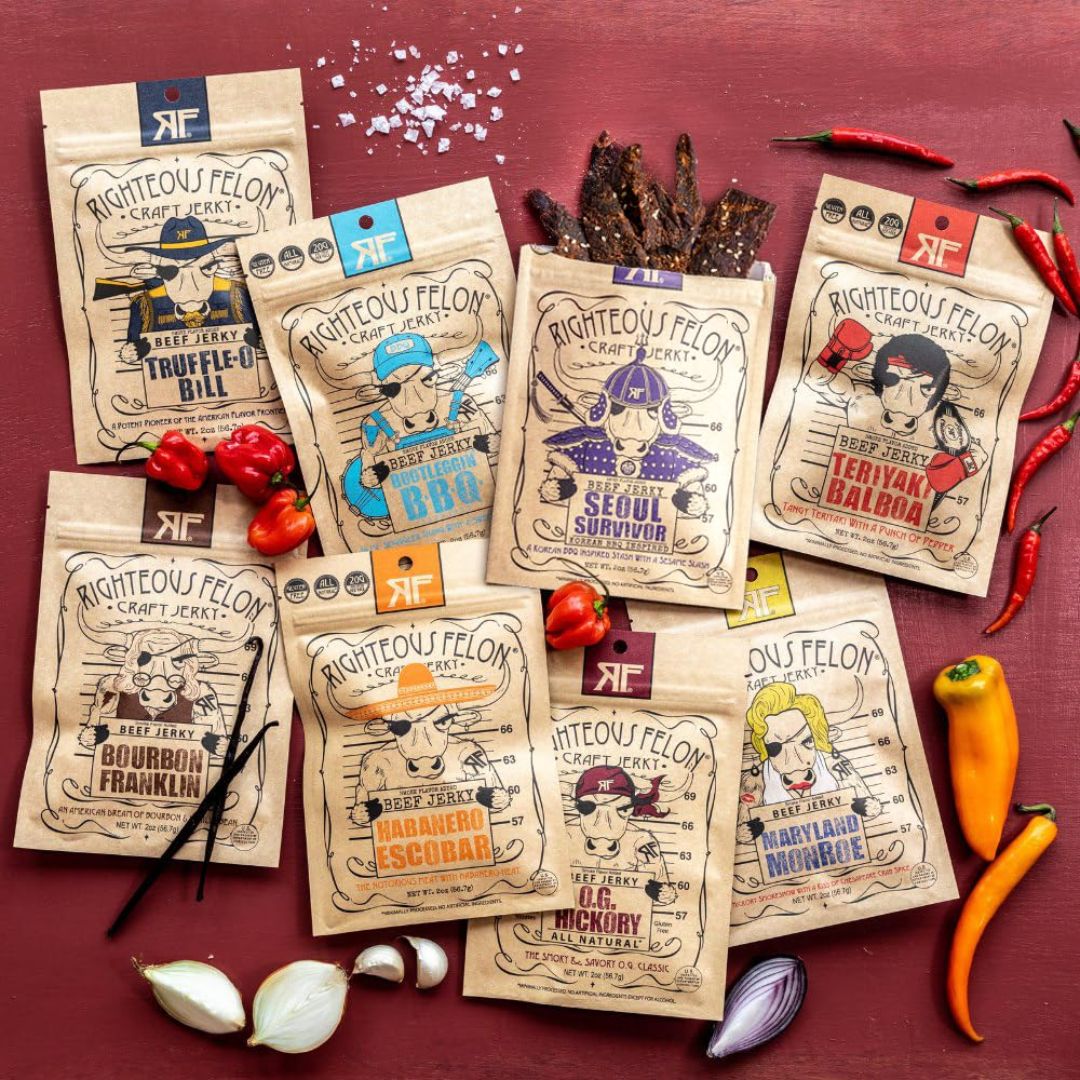 Eight bags of Righteous Felon beef jerky arranged on a dark red surface with food props including tomatoes, garlic, onions, peppers, and vanilla beans. The bags feature different illustrated character designs and flavor names such as Truffule Bill, Seoul Survivor, and Bourbon Frankin.