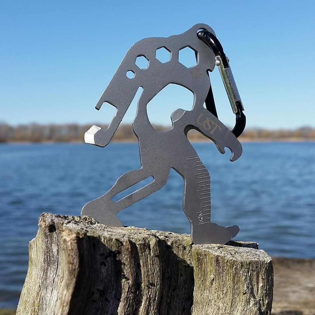 A metal Bigfoot shaped multi-tool keychain, attached to a black carabiner, standing on a weathered wooden post with a lake and blue sky in the background.