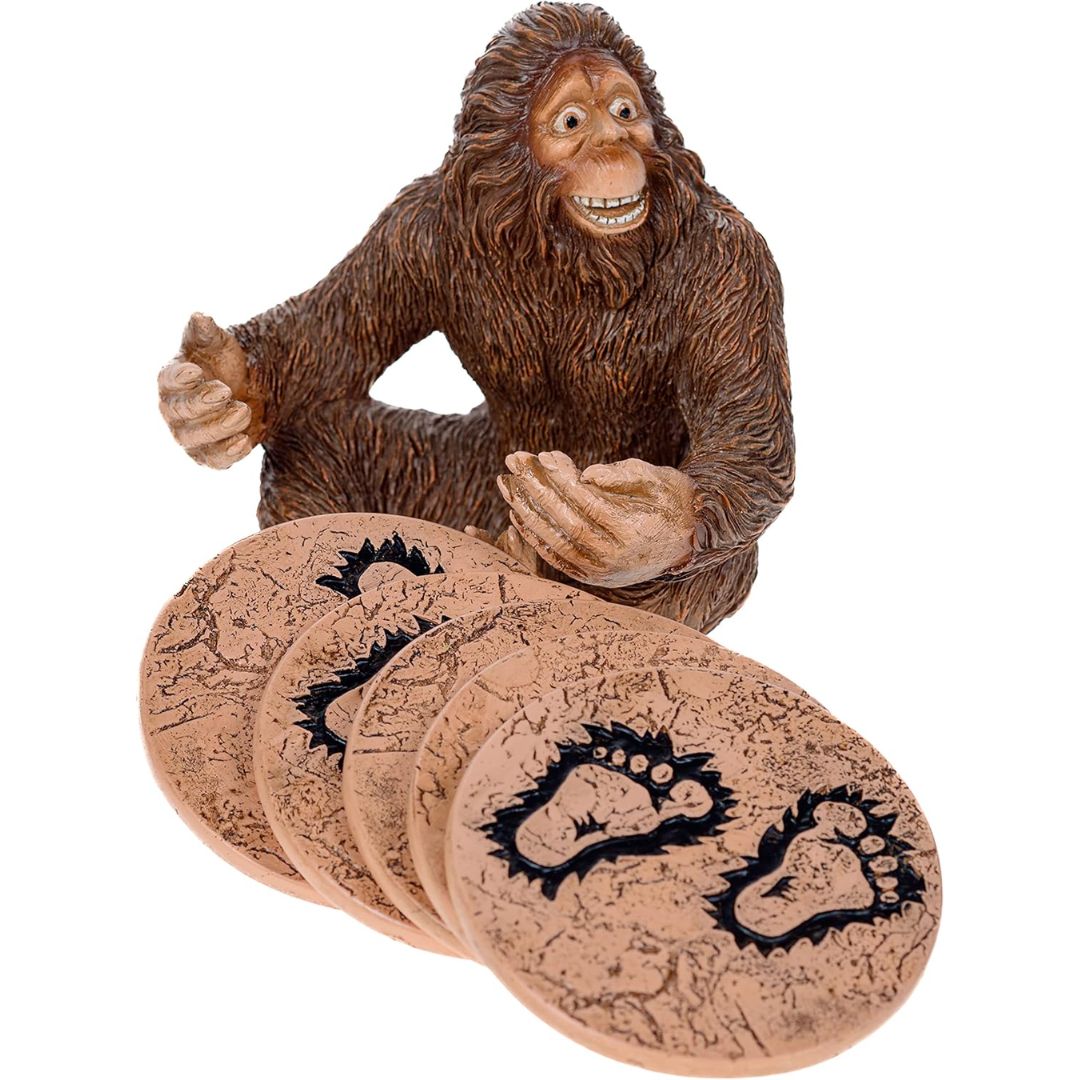 A brown furry Bigfoot figurine seated behind a fan of oval tan coasters printed with black footprint designs, shown on a plain white background.