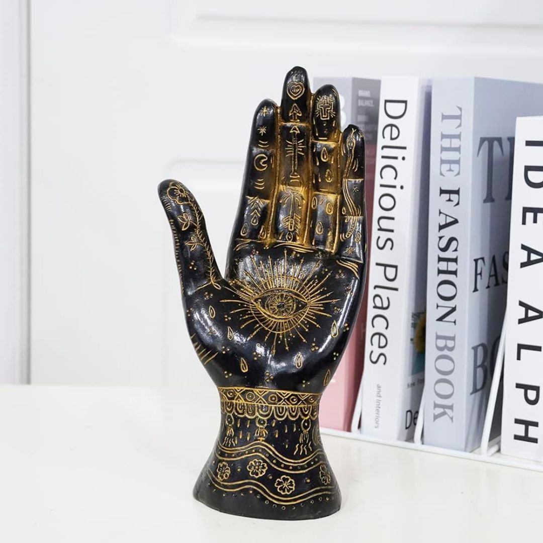 A glossy black decorative palmisty hand sculpture with intricate gold henna-style patterns and an eye motif on the palm, displayed on a white surface in front of a row of books.