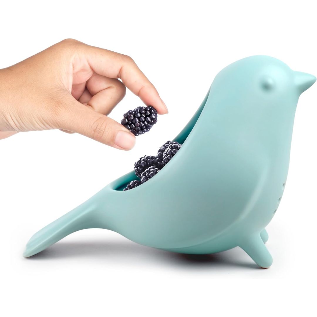 Light blue ceramic blue bird colander with a hand placing blackberries inside on a white background
