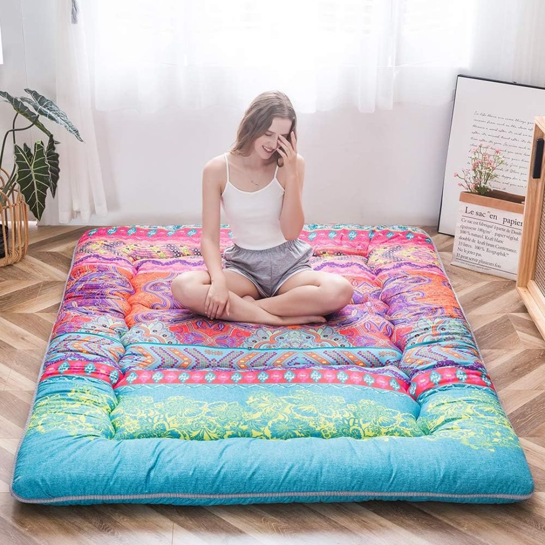 Person sitting on a colorful japanese floor mattress in a room with a plant and framed picture.