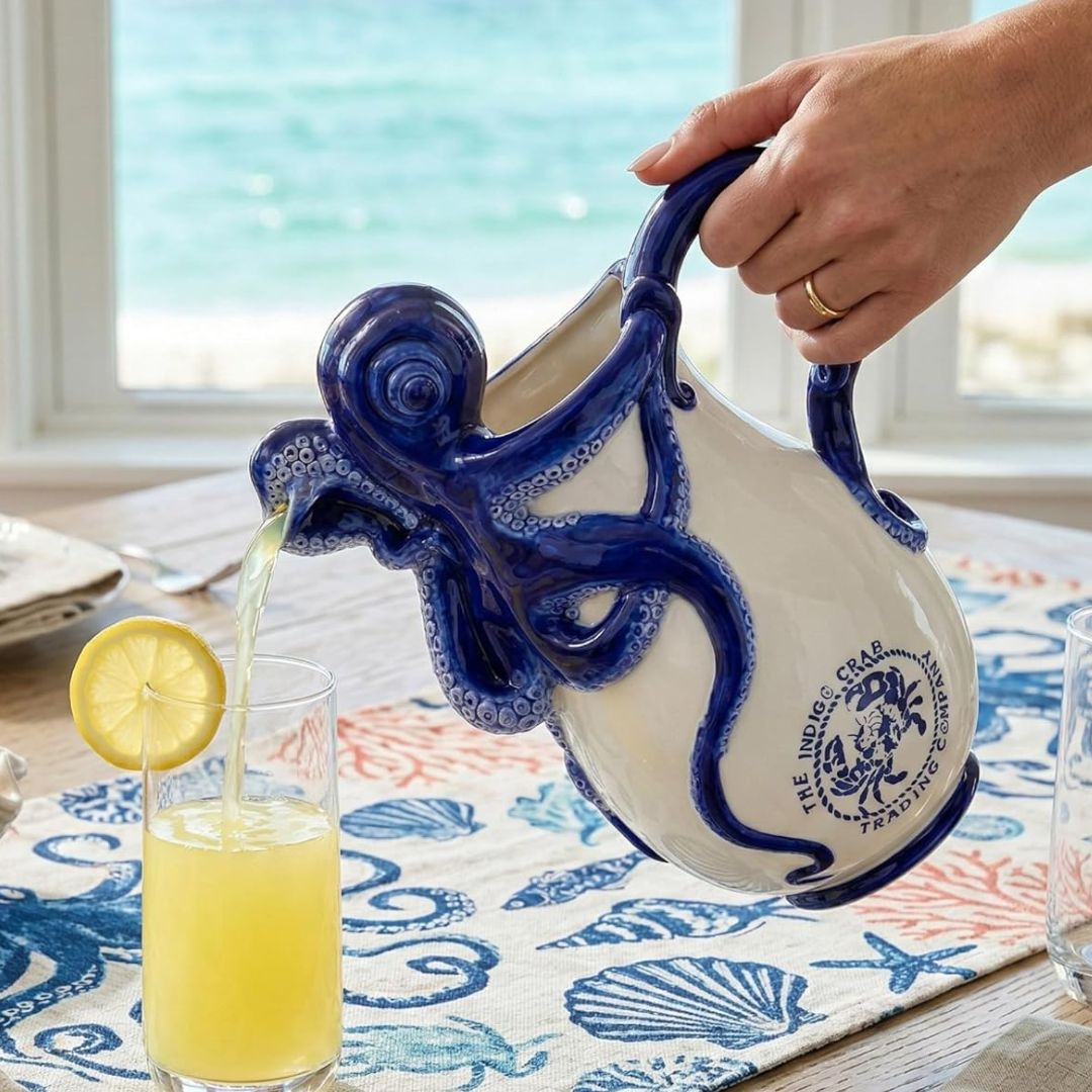 A hand holds a ceramic octopus-shaped pitcher with blue and white glaze, pouring yellow beverage into a glass with a lemon slice, set on a table by the beach.