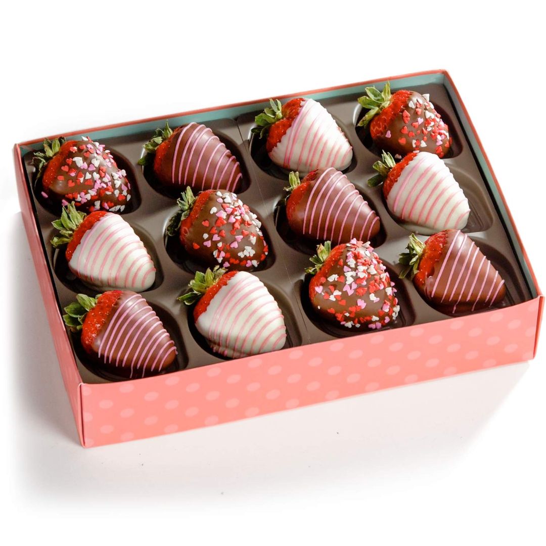 A box of 12 chocolate-dipped strawberries arranged in a pink gift box with white polka dots. The strawberries are dipped in dark and milk chocolate, some decorated with white drizzle, green leaf tops, or red and white sprinkles.