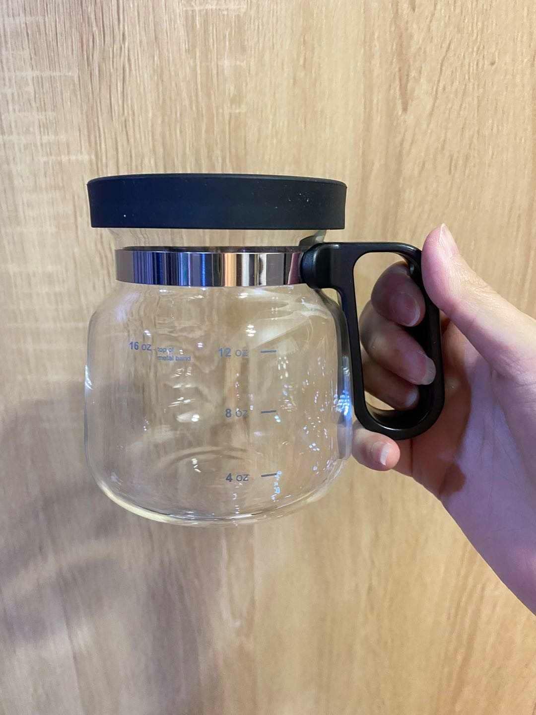 Clear glass mug that looks like a coffee carafe with black lid and handle held by a hand on a wooden surface