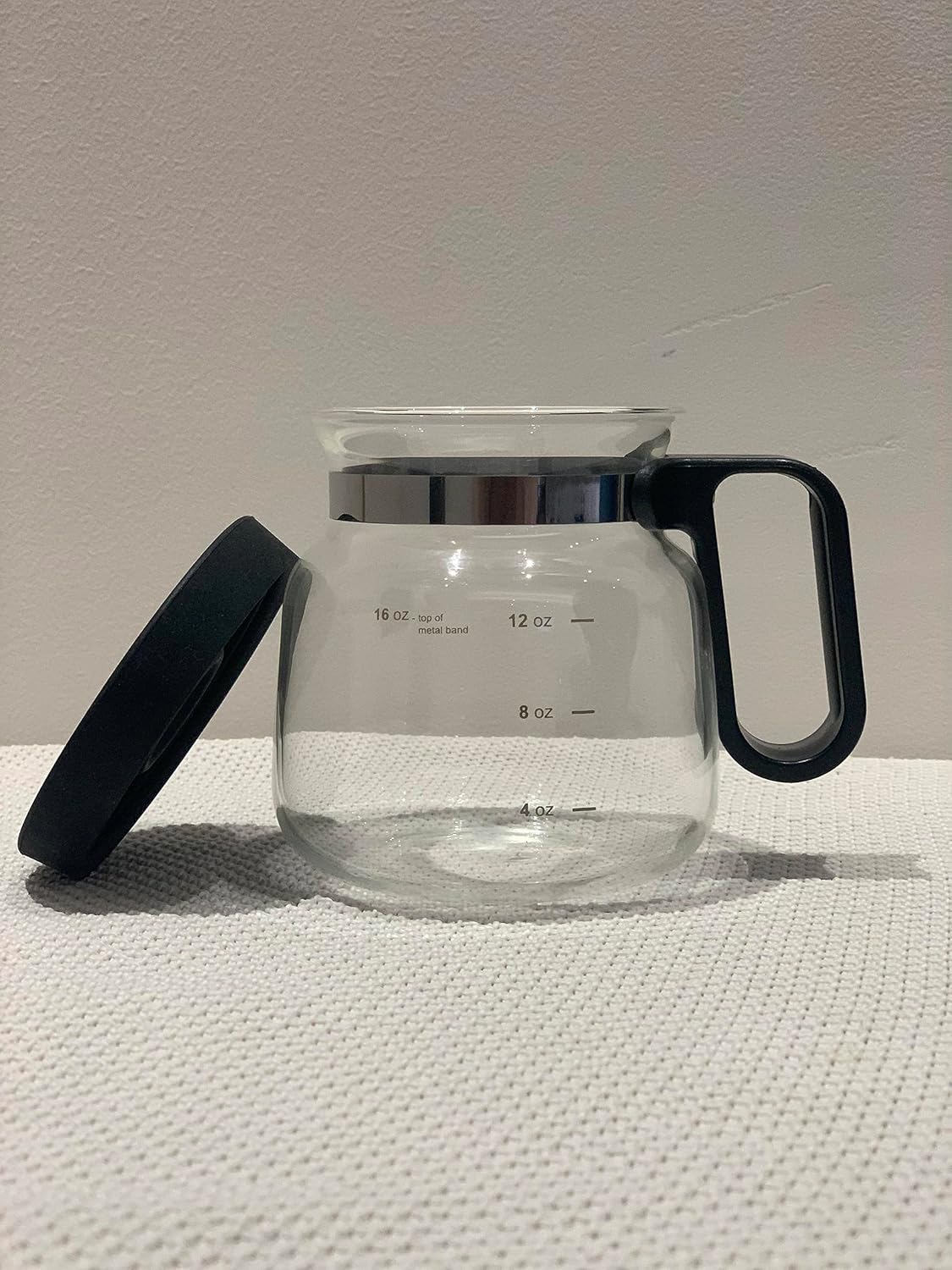 Clear glass coffee mug that looks like a coffee carafe with black handle and lid on a textured surface