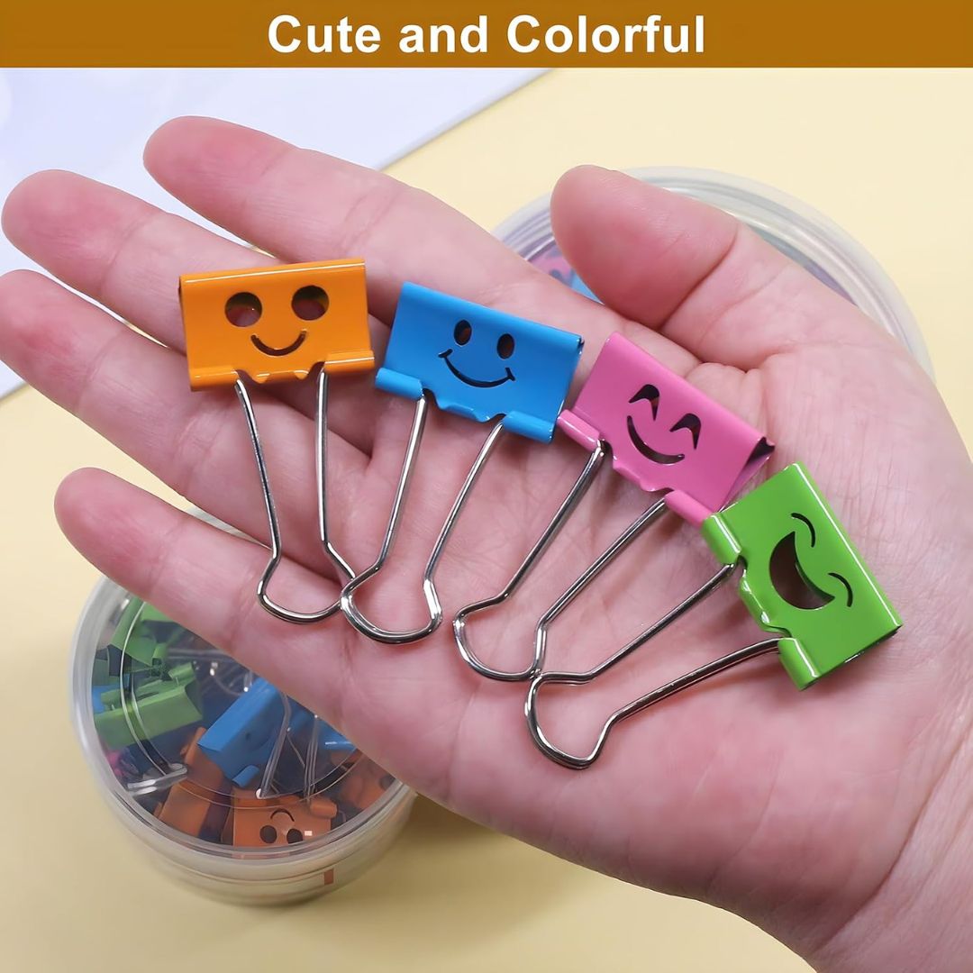 Four small metal paperclip-style binder clips with smiley face cutouts in orange, blue, pink, and green, held in a person's hand. A clear round container of more clips is visible in the background, with the text 'Cute and Colorful' at the top.