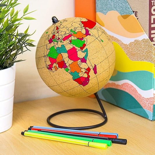 Cork globe with colorful world map on a wooden surface next to markers and a plant.