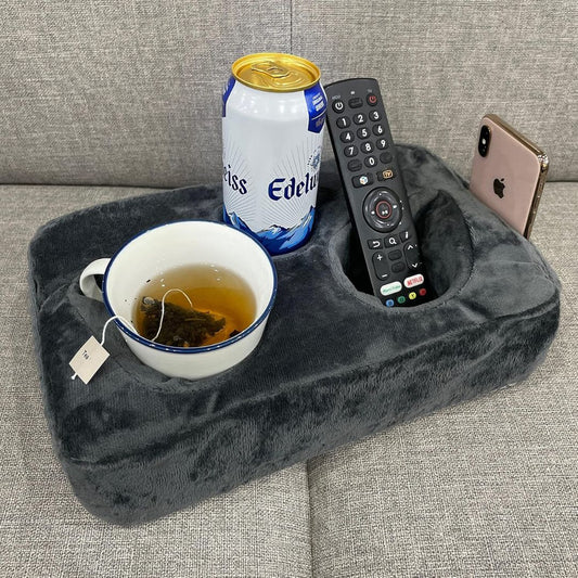 A dark gray plush tea-time tray pillow caddy resting on a gray fabric sofa, holding a white teacup with tea, a can of Edelweiss, a black TV remote, and a smartphone.