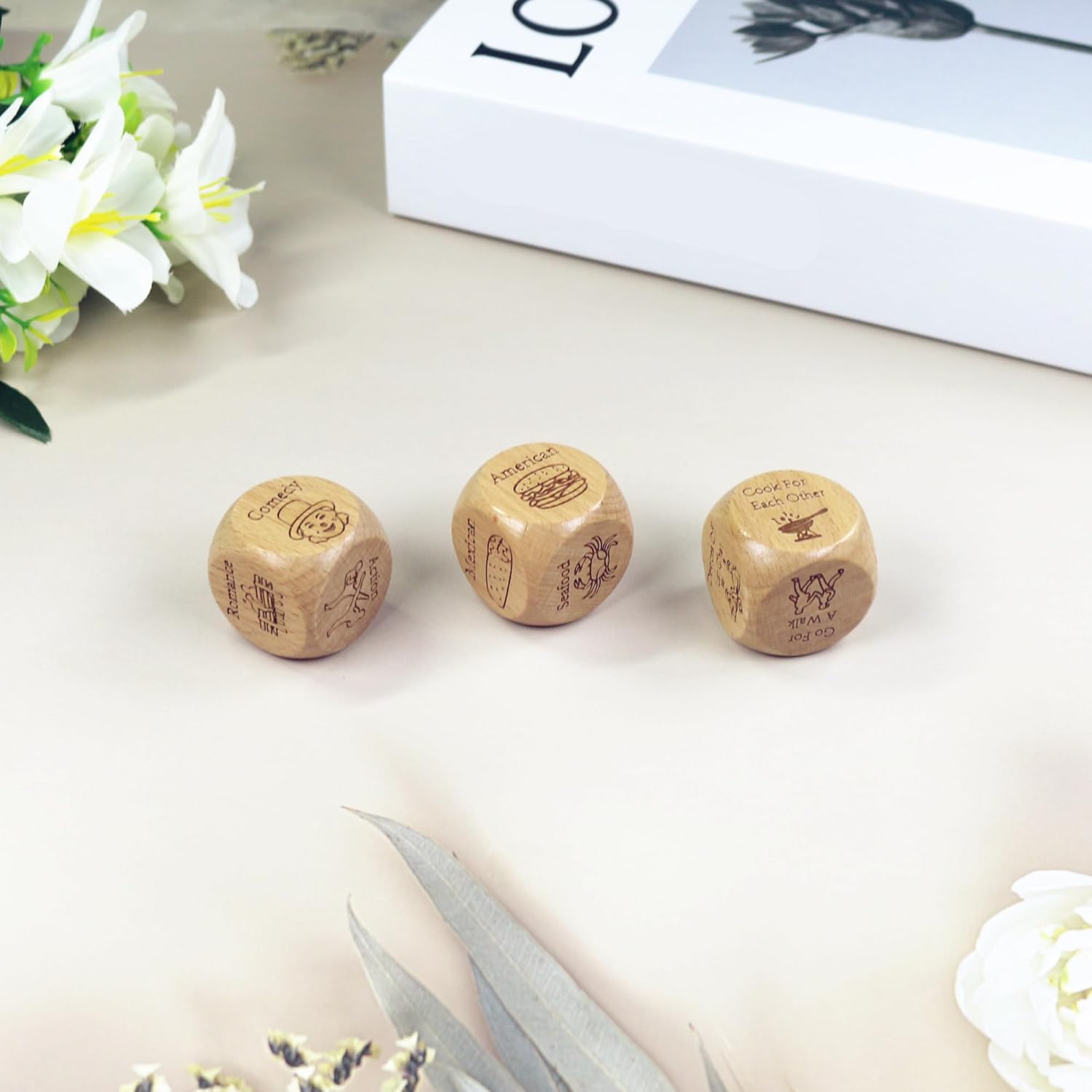 Three wooden couples date night dice with engraved designs on a light surface with flowers and a book in the background.