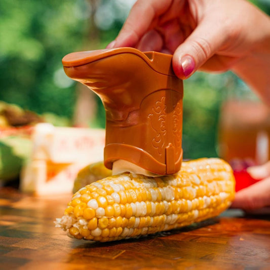 Boot shaped butter spreader being used on a corn cob with a blurred outdoor background