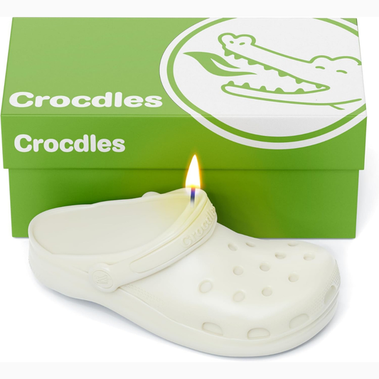 White croc-shaped candle with a lit wick in front of a green box labeled 'Croccles'.