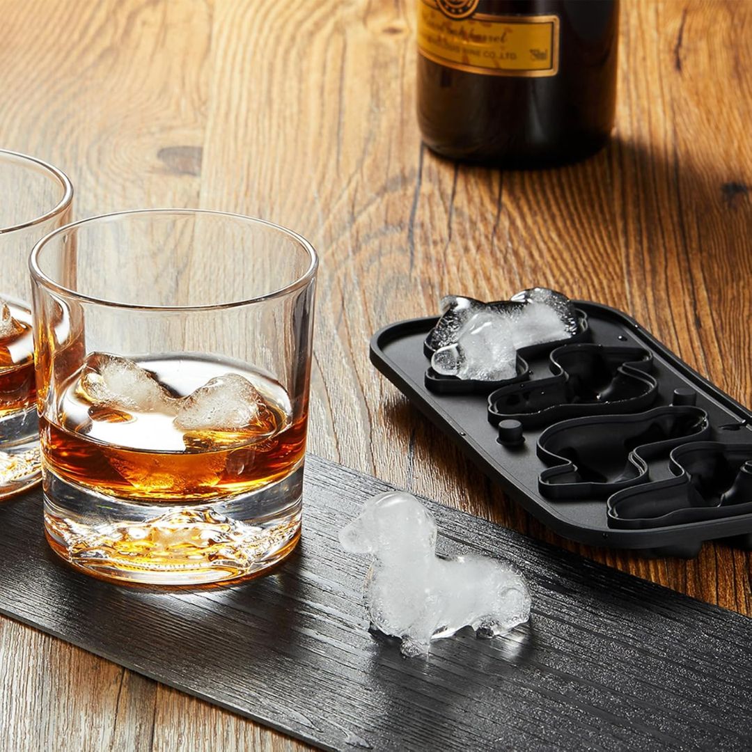 A black silicone dachshund-shaped ice cube mold tray with a dachshund-shaped ice cube sits on a wooden table beside two whiskey glasses with ice, along with loose ice cubes and a bottle in the background.