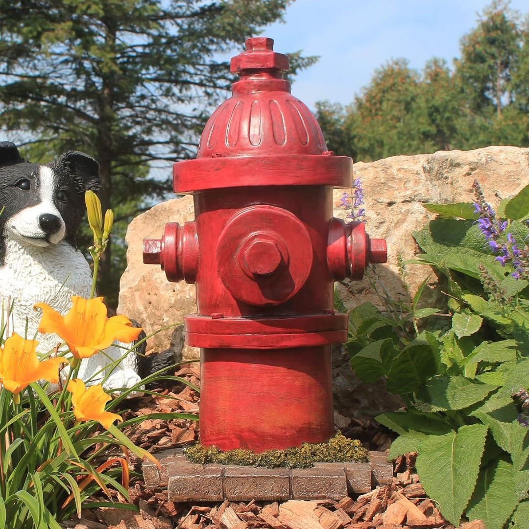 A red fire hydrant garden statue placed outdoors on a brick base among mulch, green plants, purple flowers, and a dog statue.
