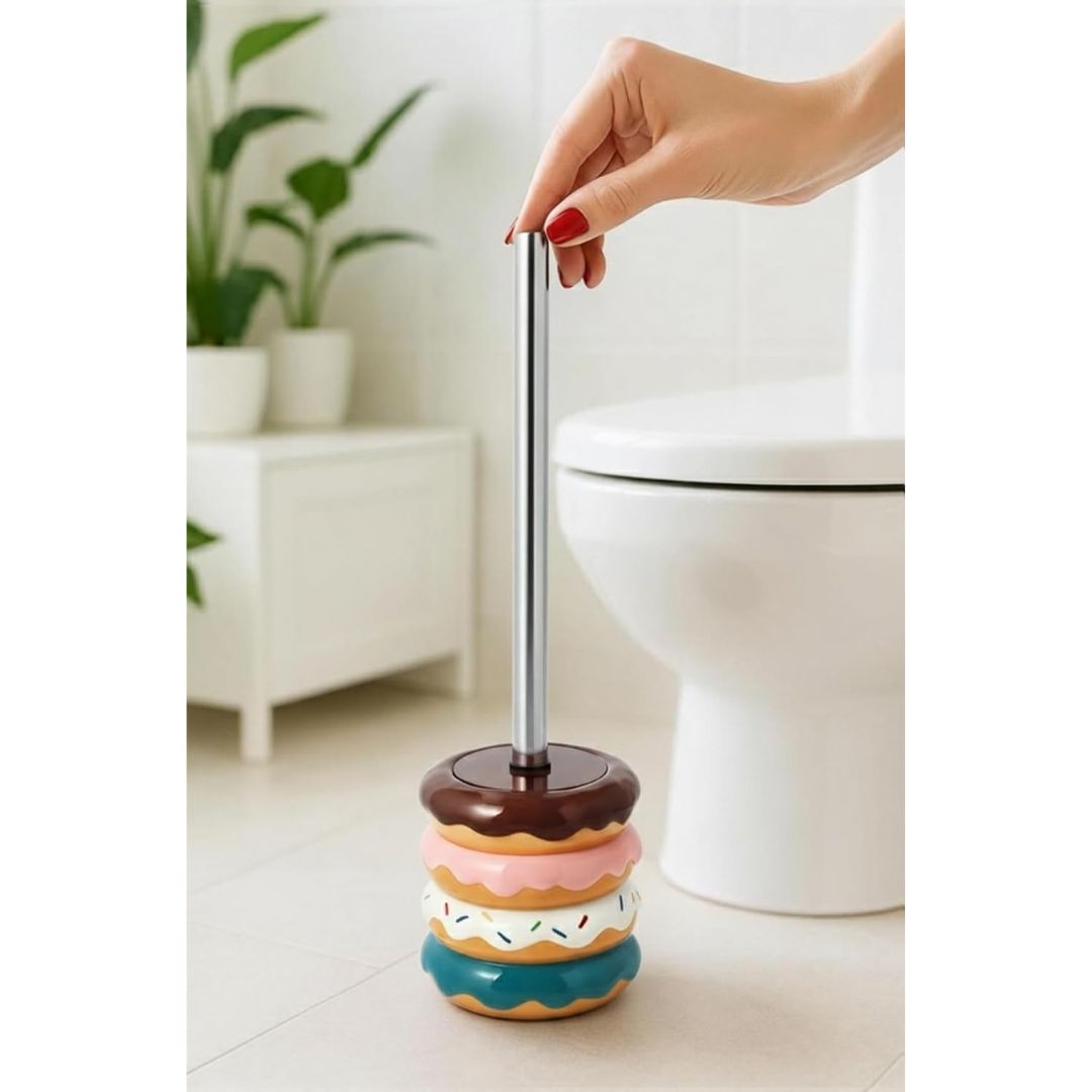 A hand with red nail polish is shown holding the top of a novelty toilet brush designed to look like stacked donuts in a bathroom setting with a white toilet and plant in the background.