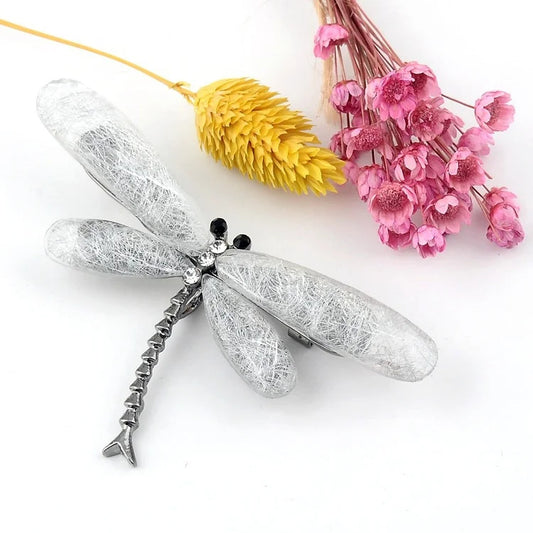 A silver dragonfly brooch with gemstone wings, adorned with black and clear gems, placed on a white surface with dried yellow foliage and pink flowers in the background.