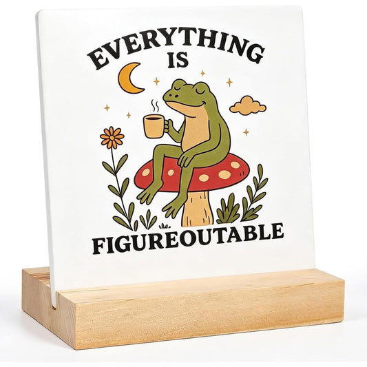 A square white decorative sign on a light wood display stand, featuring a whimsical illustration of a green frog sitting on a red mushroom and holding a mug, with the text 'EVERYTHING IS FIGUREOUTABLE'.