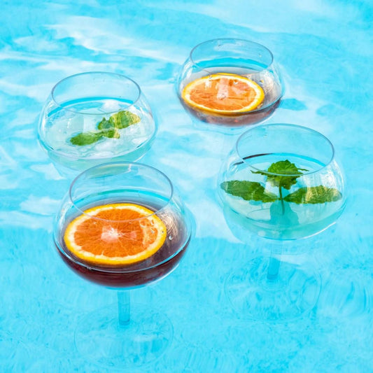 Four floating shatterproof wine glasses with orange slices and mint leaves floating in a pool.
