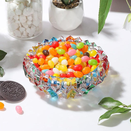 Colorful jelly beans in a decorative glass bowl on a light surface with a cookie and plant in the background.