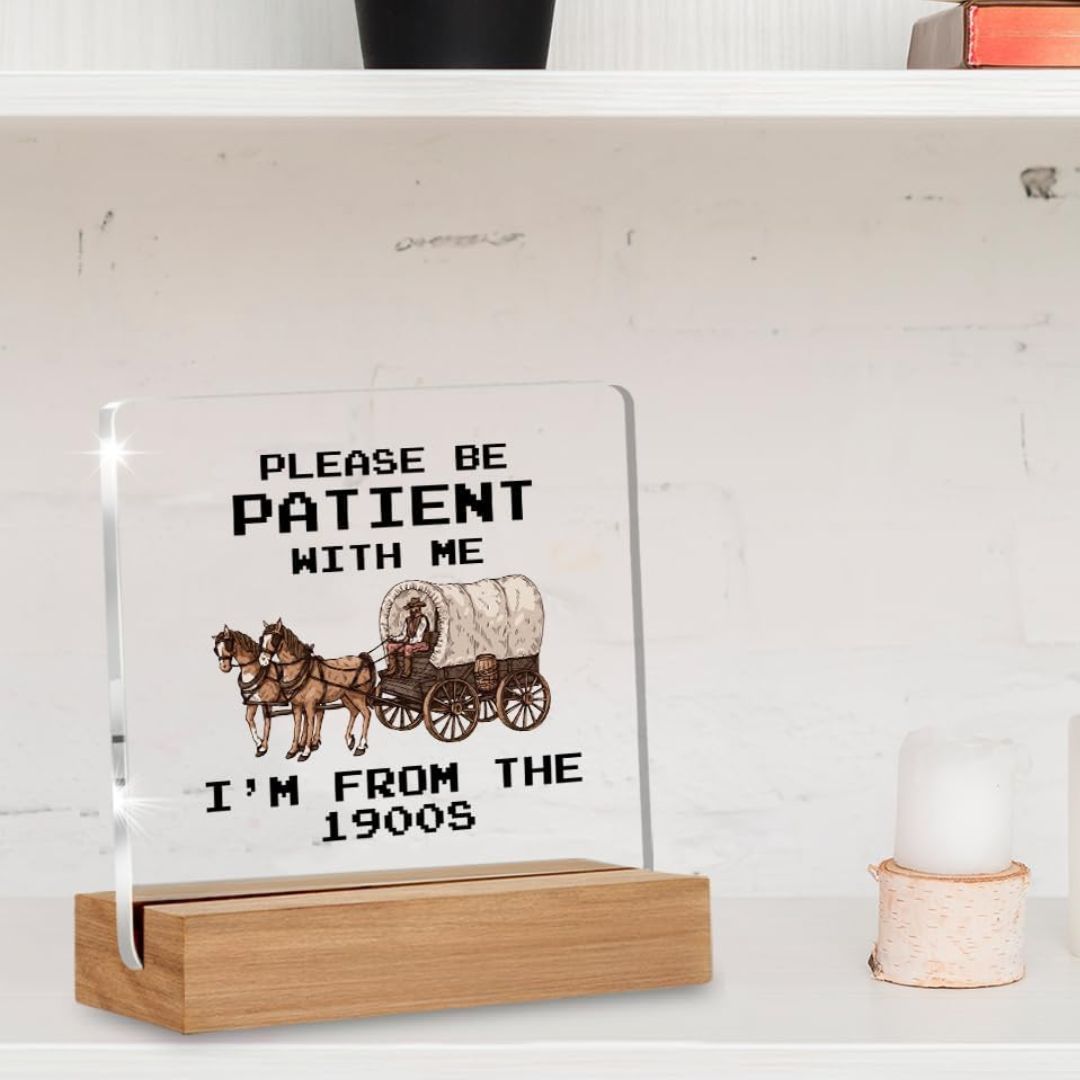 A funny glass and wood sign with text reading 'Please be patient with me I'm not the 1900s' on a shelf.