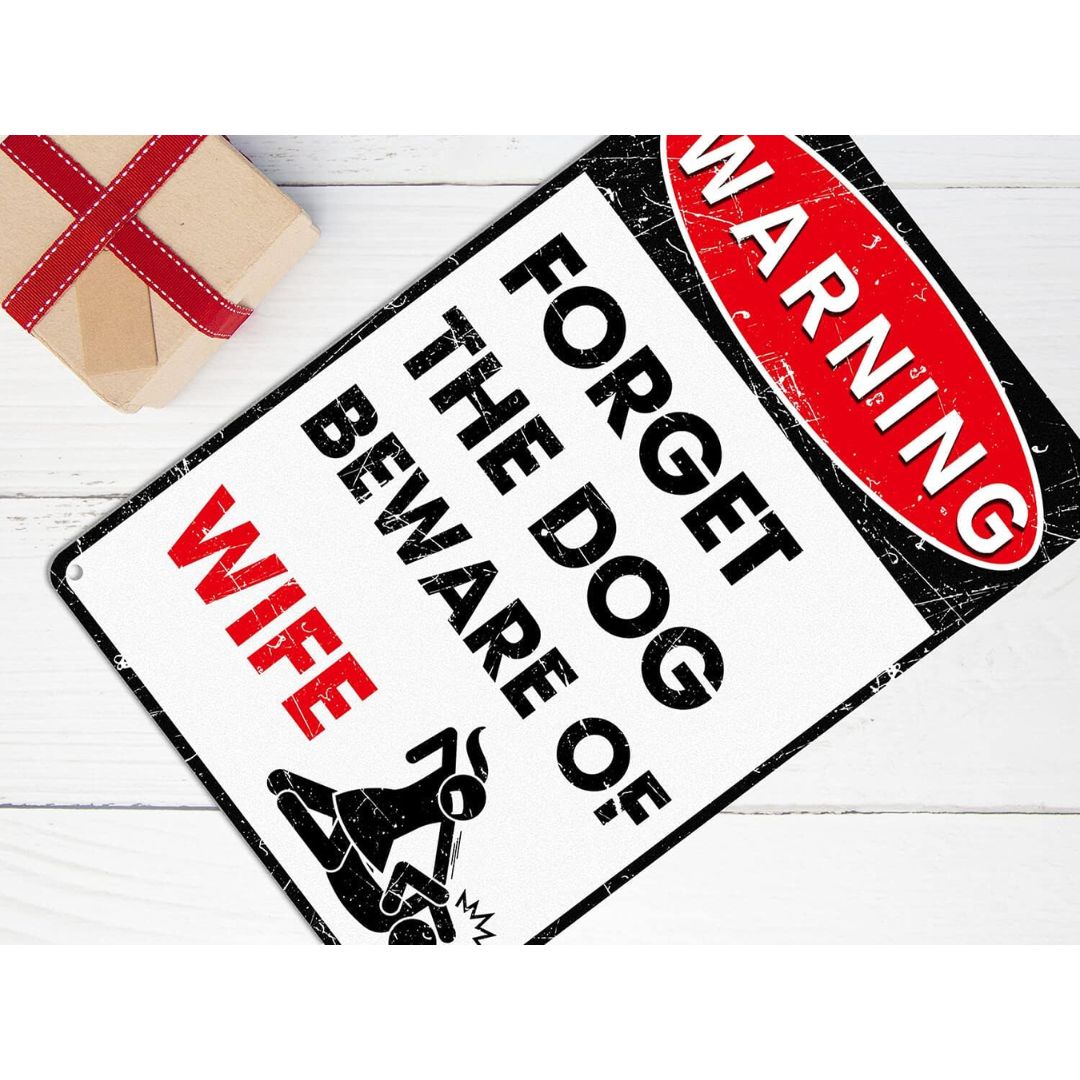 Funny metal warning sign reading “Forget the dog beware of wife” displayed on a wooden surface with a gift box