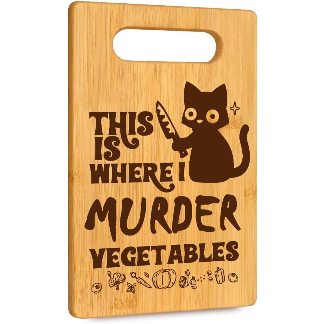 A rectangular bamboo cutting board with a handle cutout at the top, featuring a dark brown engraved design of a cartoon cat holding a knife and the text 'THIS IS WHERE I MURDER VEGETABLES' along with small vegetable icons.